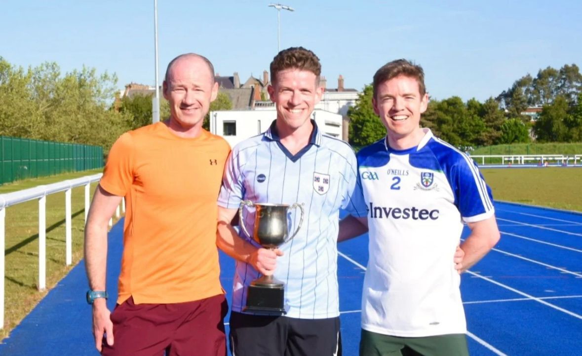 Three runners on an athletics track, the one in the middle holding the CAT 5K Fun Run trophy